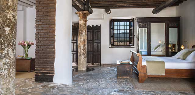 Schlafzimmer der Familiensuite Los Aljibes mit Steinsäulen, die eine Holzbalkendecke stützen.